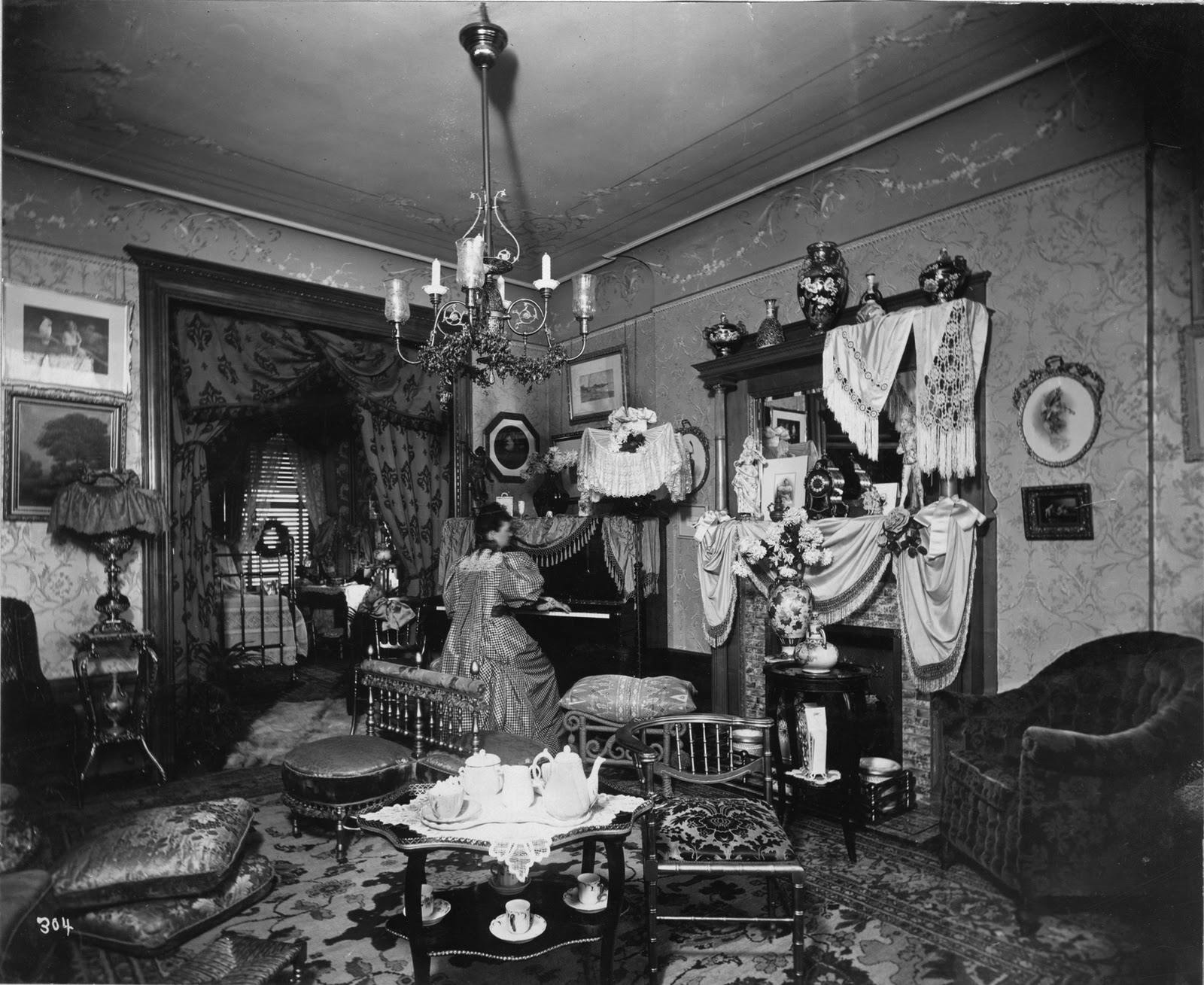 Black and white photograph of a crowded living room. A woman is playing the piano in the background